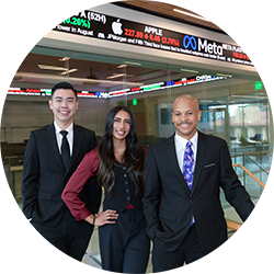 Three smiling graduate students from the UCR School of Business standing in front of a stock market ticker display. front of 