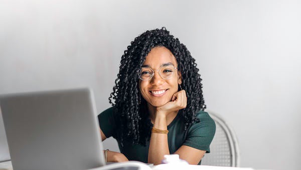 young woman smiling, sitting at laptop, chin on hand